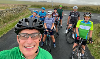 A group of eight cyclists, stopped on the road, surrounded by green fields, with some trees in the distance 