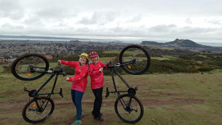 Leanne and Jenn, another Breeze Champion MTB leader, on a hill overlooking Edinburgh, posing with their bikes by lifting the front wheels into the air