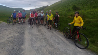 Picture of a group of 14 cyclists standing with their bikes on a gravel track, surrounded by green hills. In the distance on the right of the image is a small wood of trees