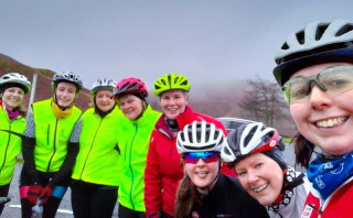 A group of 8 female cyclists posing for the camera, all are smiling, despite the sky looking overcast and grey