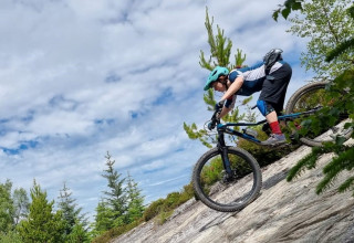 Leanne on a mountain bike, cycling in the attack position going downhill on a rock, surrounded by trees and cloudy blue sky 